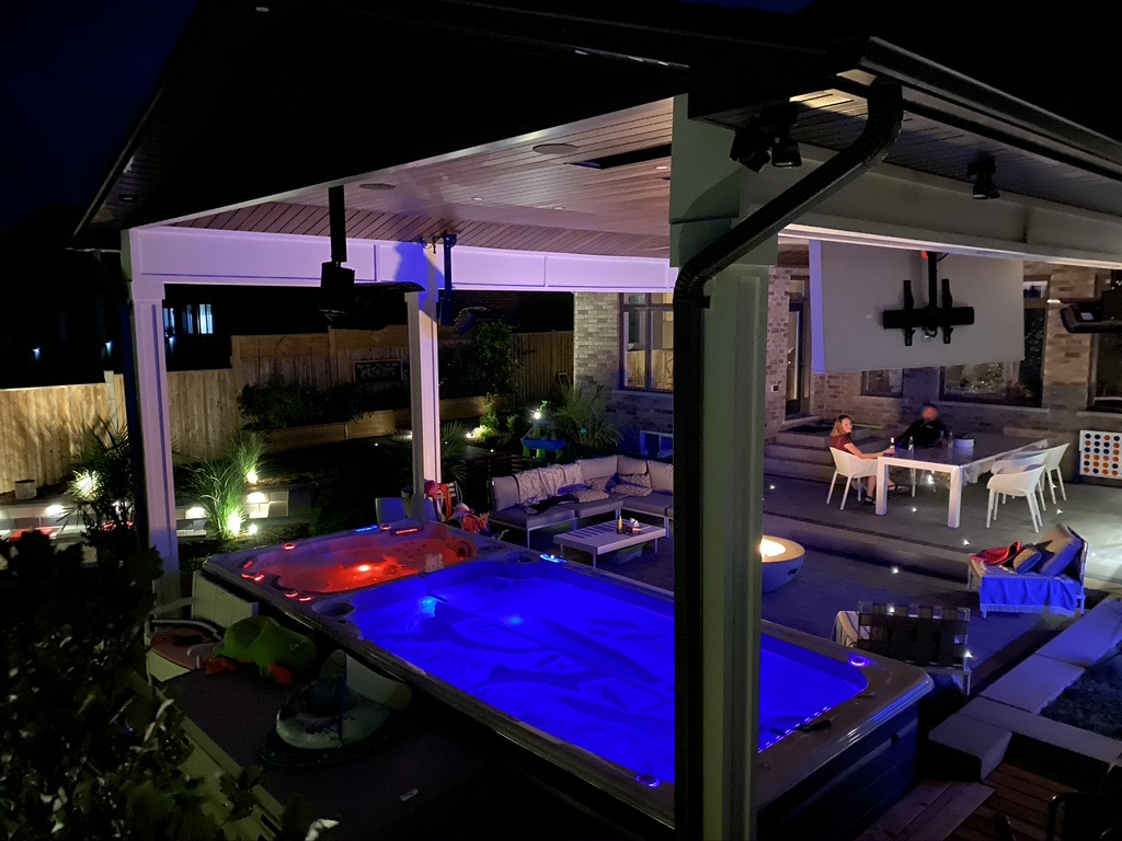 Illuminated backyard at night with covered cabana, swim spa, fire feature and outdoor lounge