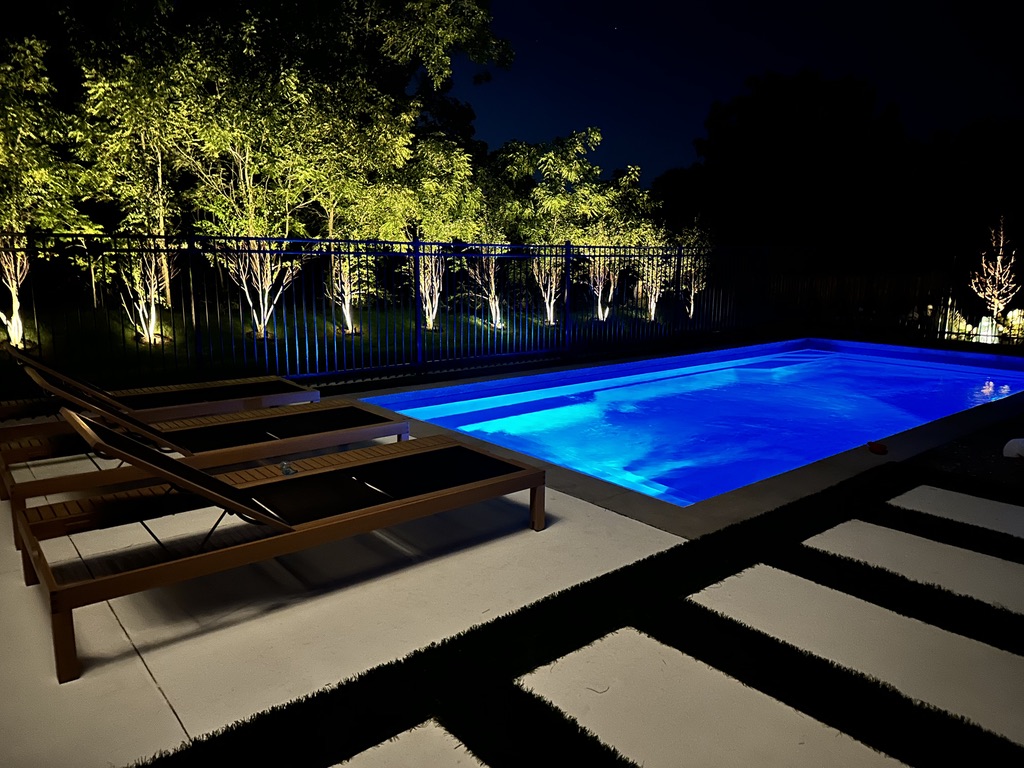 Fiberglass pool glowing blue at night with uplit landscaping
